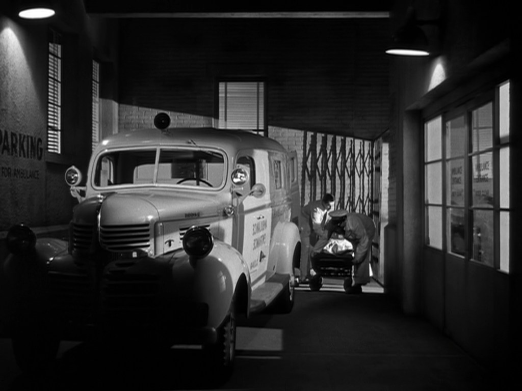 A black and white image depicting an ambulance parked in a dimly lit garage, with two people in medical attire attending to a stretcher.