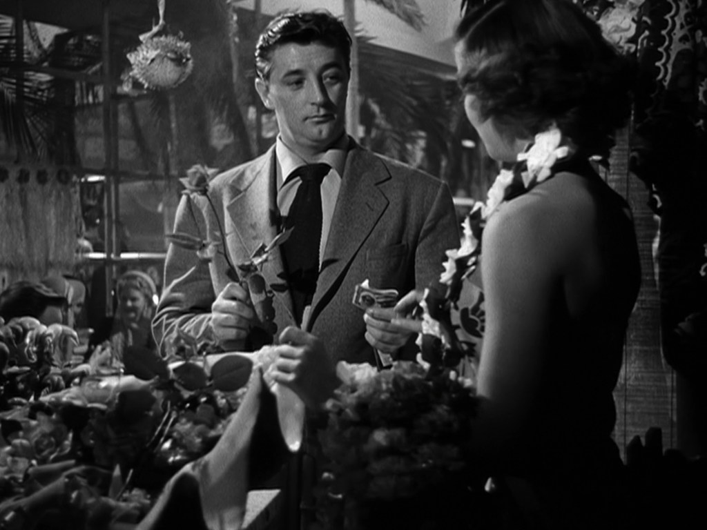 A man in a suit holding flowers and cash engages in conversation with a woman wearing a flower lei, surrounded by colorful decorations in a lively setting.