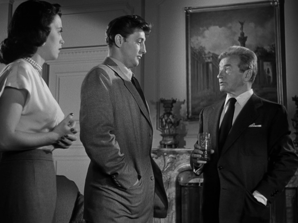 A black and white scene featuring three characters: a woman in a white blouse, a man in a gray suit, and another man in a black suit holding a glass. They are engaged in a conversation, with a portrait and ornate decor in the background.