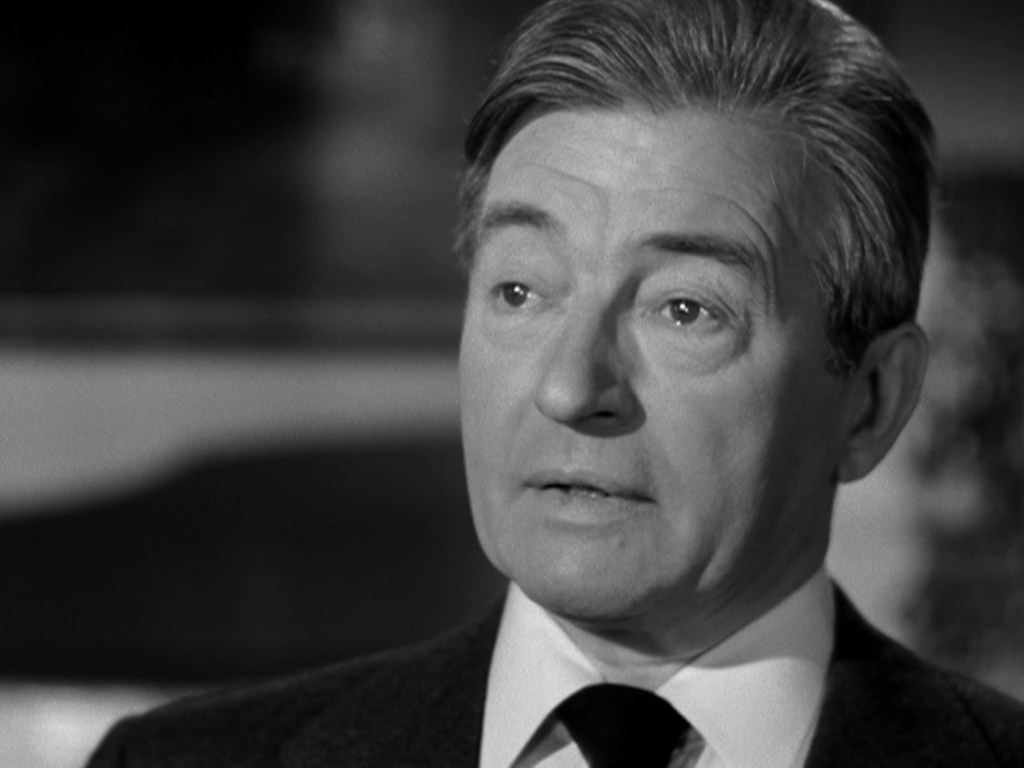 Close-up of a man in a suit and tie, with silver hair, appearing thoughtful or concerned in a black and white setting.