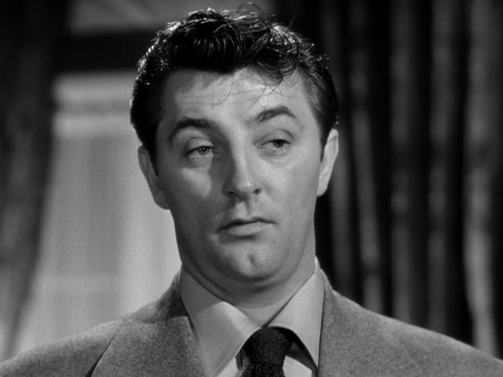 Close-up of a man with dark, slicked-back hair, wearing a grey suit and tie, looking thoughtfully with a slight expression of skepticism.