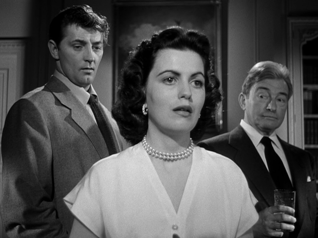 A woman in a white blouse with pearl necklace stands in focus, flanked by two men in suits, one looking concerned and the other holding a glass and appearing intrigued.