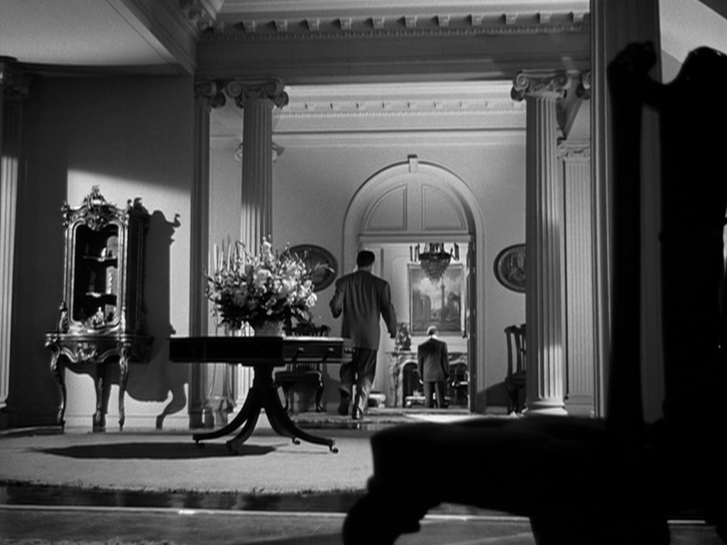 A black and white interior shot of a grand room, featuring ornate decor including a floral arrangement, a decorative cabinet, and large columns. Two men are visible, one walking towards the background and another sitting down. The composition suggests a sense of depth and elegance.