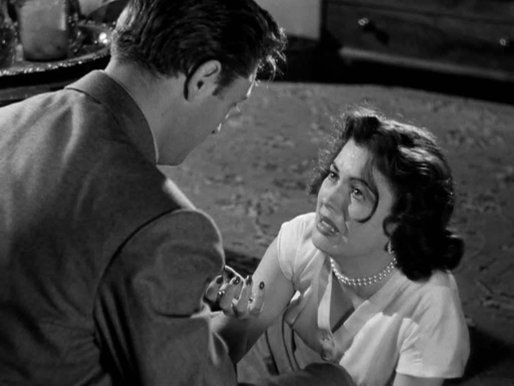 A dramatic scene from a classic black-and-white film, featuring a man in a suit leaning towards a woman on the floor who appears distressed, wearing a white blouse and a pearl necklace.