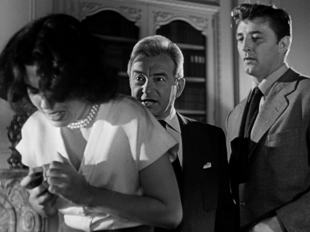 A tense scene featuring a woman in distress, holding her hands tightly, while a man with a stern expression confronts her, and another man watches with concern.