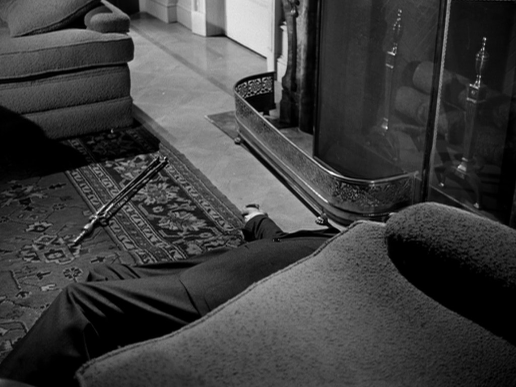 A man lying on the floor in a living room, partially obscured by a couch, with a decorative rug and a fireplace in the background.