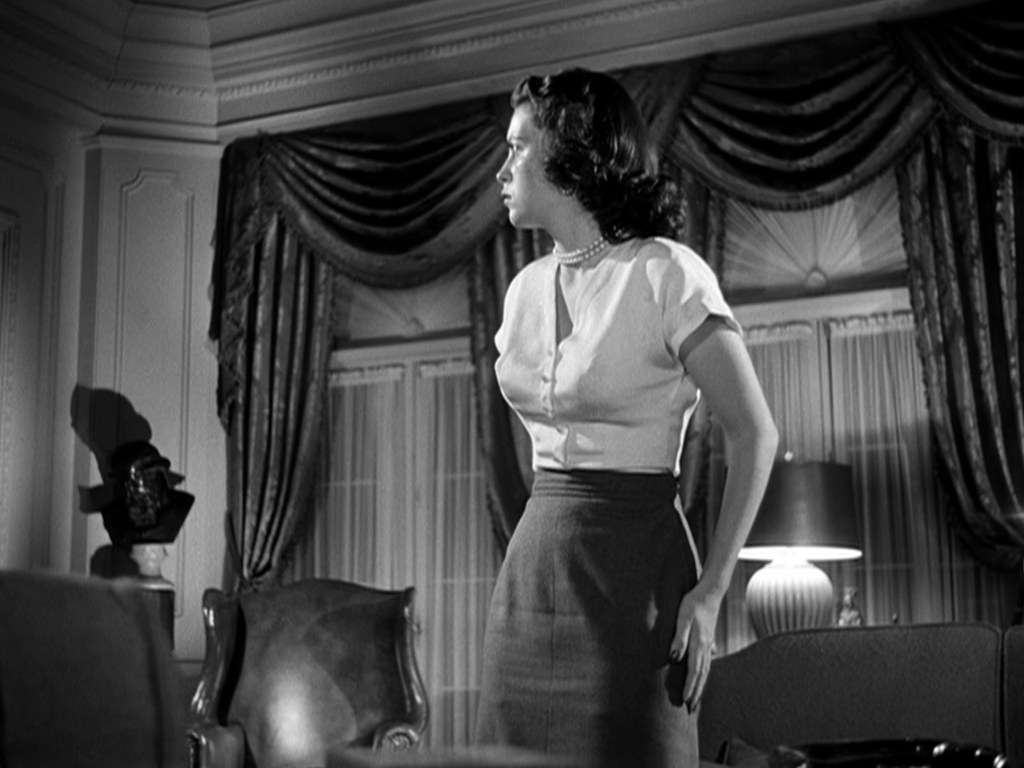 A woman with dark, wavy hair is standing in a living room, wearing a short-sleeved top and a pencil skirt. She is looking to the side with a thoughtful expression, with elegant curtains and a lamp visible in the background.