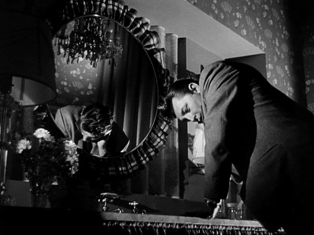 A man in a suit leans over a sink, reflected in a round mirror, with decorative wallpaper and a lamp visible in the background.