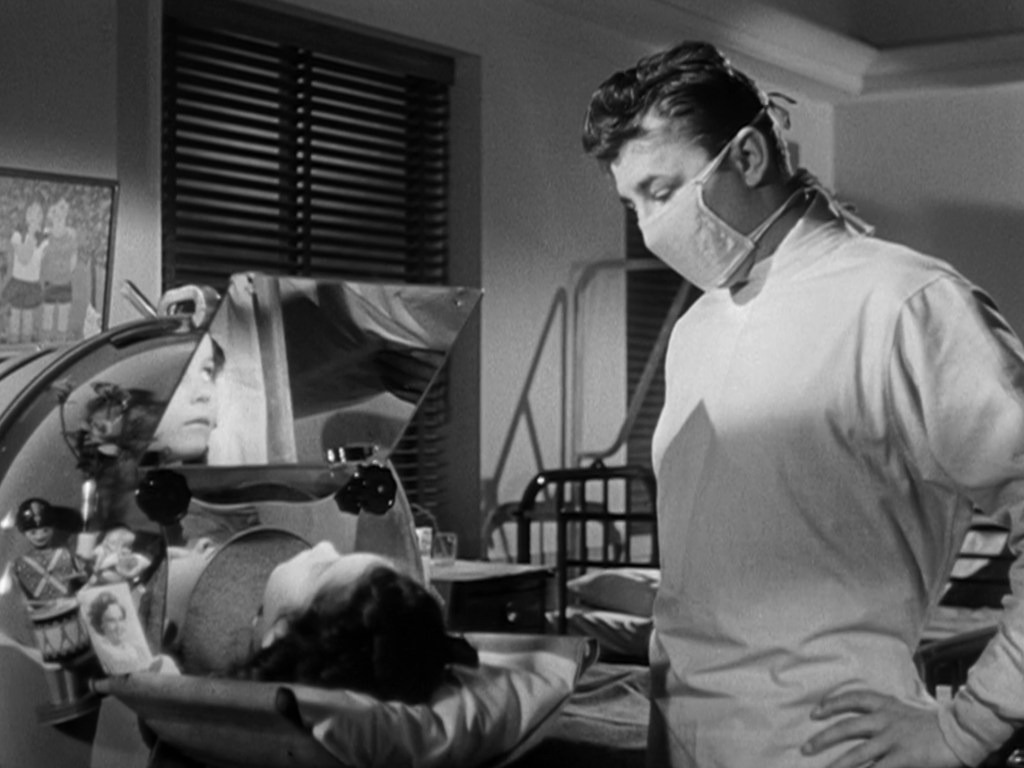 A doctor in a surgical mask and gown observes a patient lying in a medical device, featuring reflective surfaces, in a hospital room with blinds.