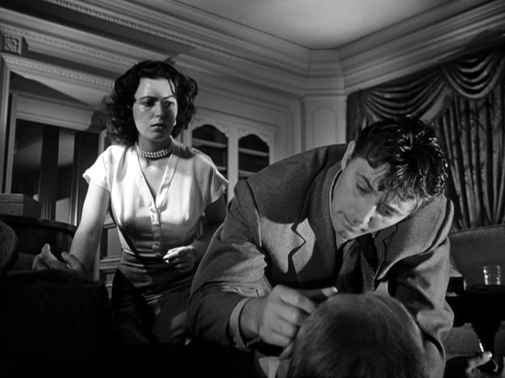A tense black and white scene featuring a man focusing intently on a man's head while a woman watches with concern in a stylishly furnished room.
