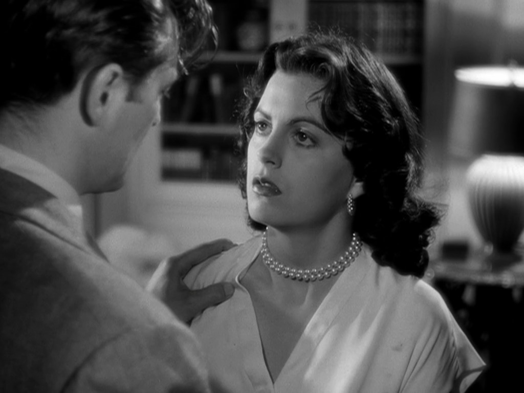 A black-and-white scene featuring a woman with dark, wavy hair wearing a pearl necklace, looking intently at a man dressed in a suit. The background includes a lamp and bookshelves.