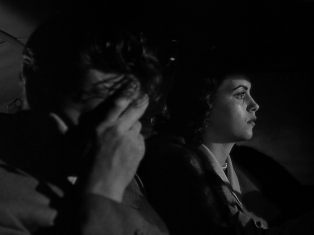 A man with his hand covering his face sits in a car next to a woman who gazes out the window, both in dim lighting.