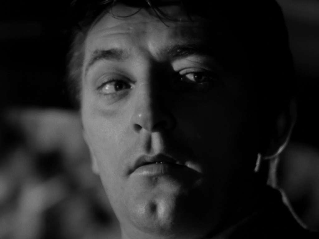 Close-up of a man in black and white, showing a serious expression with dramatic lighting highlighting his facial features.