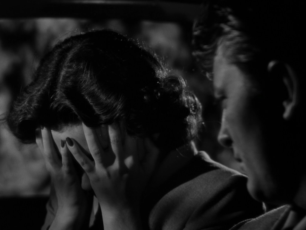 A woman with dark, curly hair sits in a car, covering her face with her hands in distress, while a man with a lighter hairstyle looks at her with concern.