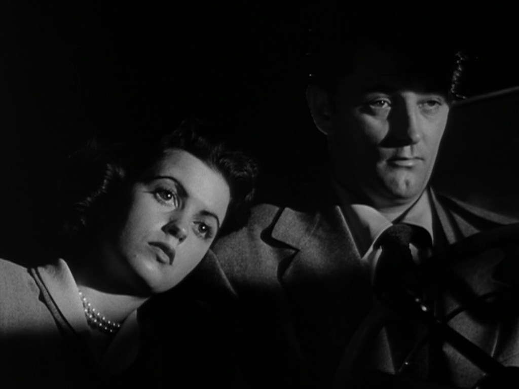 A woman rests her head on the shoulder of a man in a dark setting, conveying a sense of intimacy and contemplation.