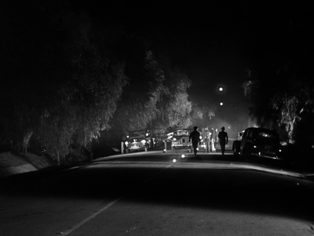 A dark street scene at night with silhouetted figures walking and parked cars along the side.