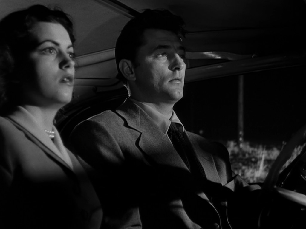 A man and a woman sitting in a car, both looking tense and concerned, with shadows highlighting their faces in a noir-style setting.