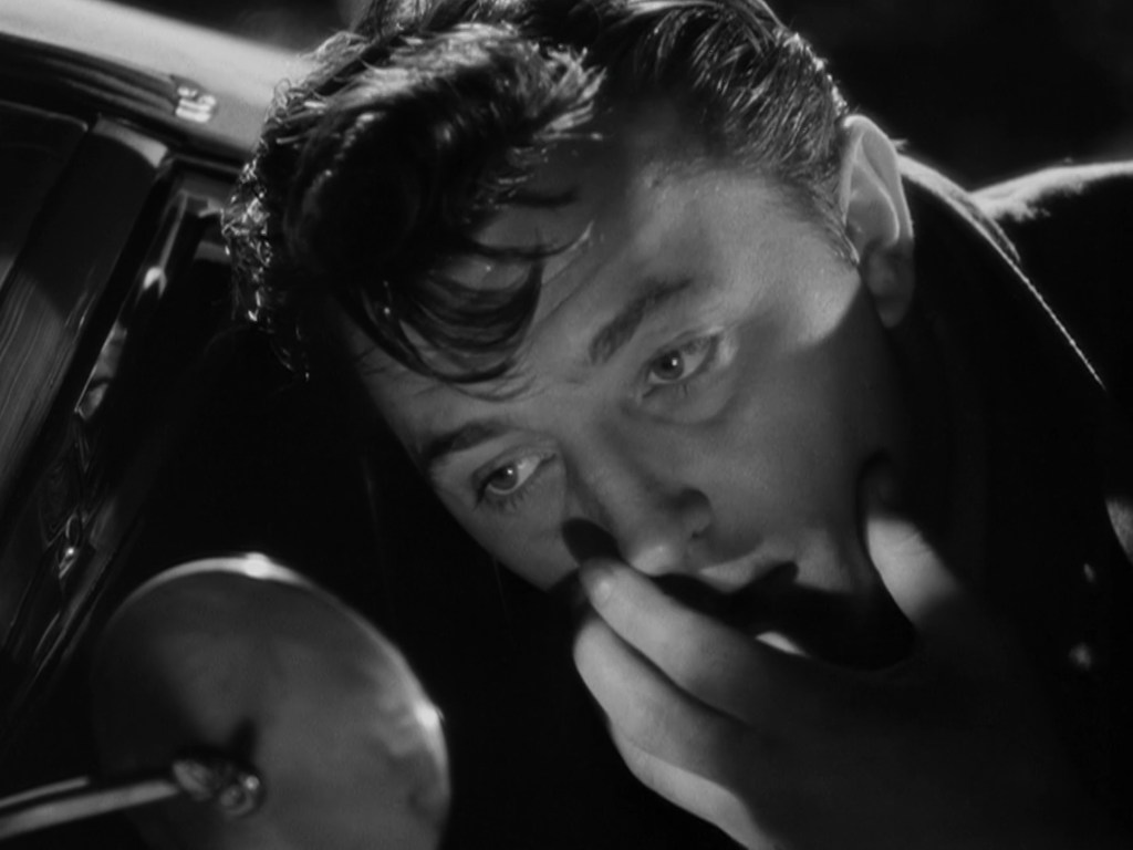 A close-up of a man looking intently into a side mirror of a car, with a thoughtful expression and his hand touching his face, in a dramatic black and white style.