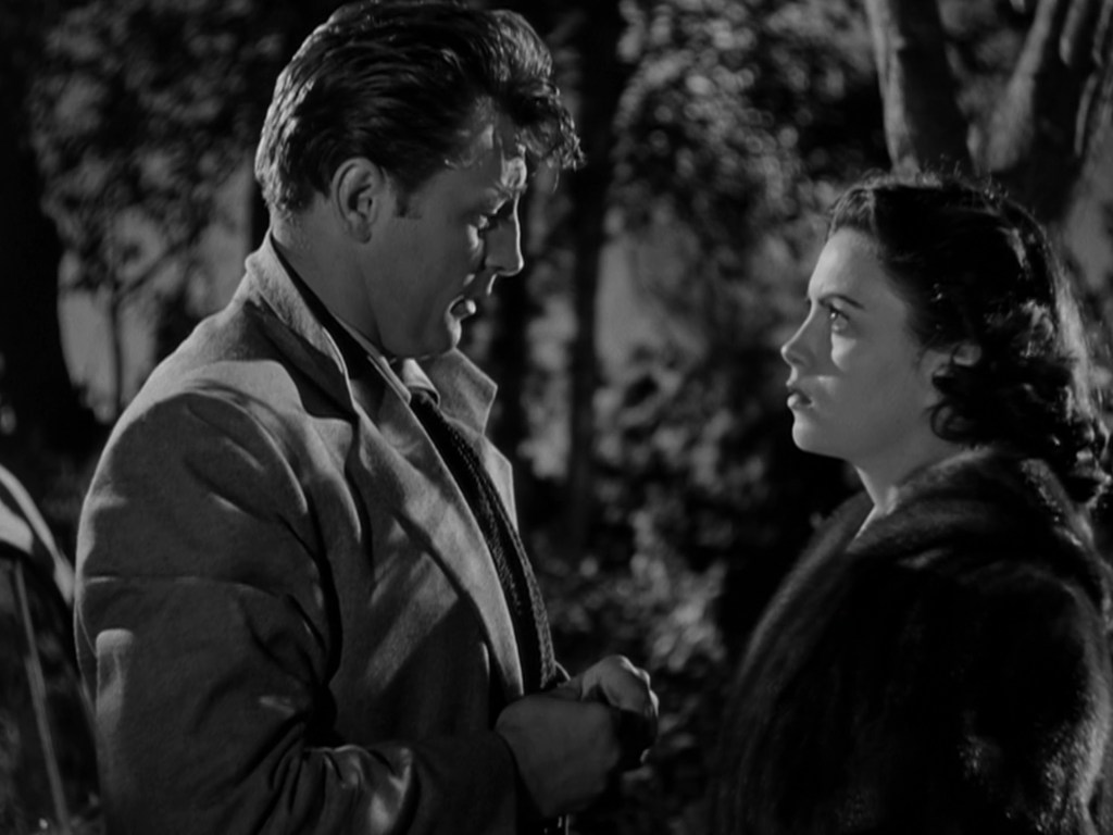 A black and white scene featuring a man and a woman in a serious conversation outdoors, surrounded by trees. The man is holding something in his hands while looking intently at the woman, who gazes back with a concerned expression.