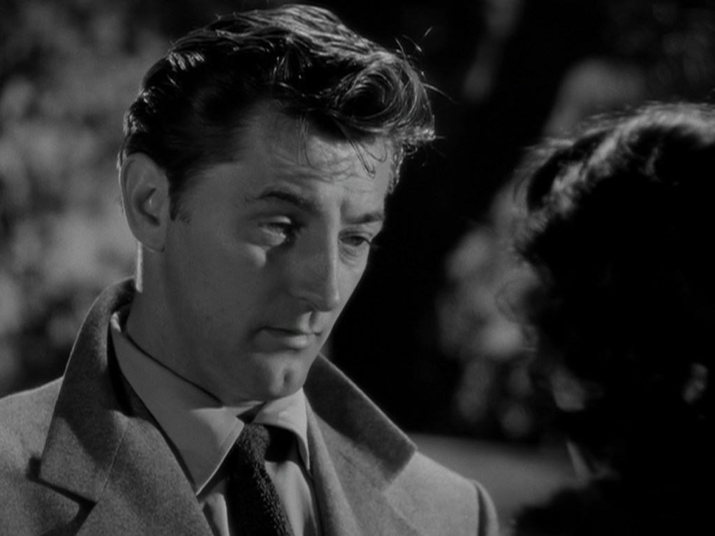 A close-up of a man with slicked-back hair wearing a gray coat and tie, looking contemplative as he engages in conversation.