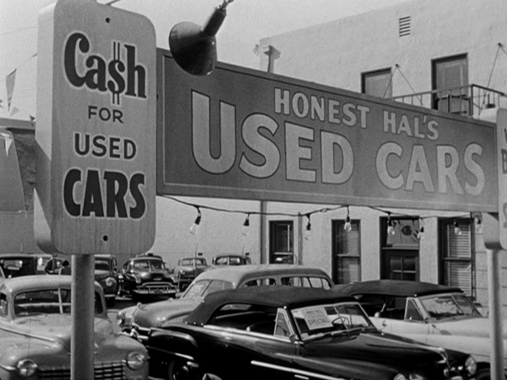 Signage for Honest Hal's Used Cars with a focus on 'Cash for Used Cars' in front of a vintage used car lot filled with parked cars.