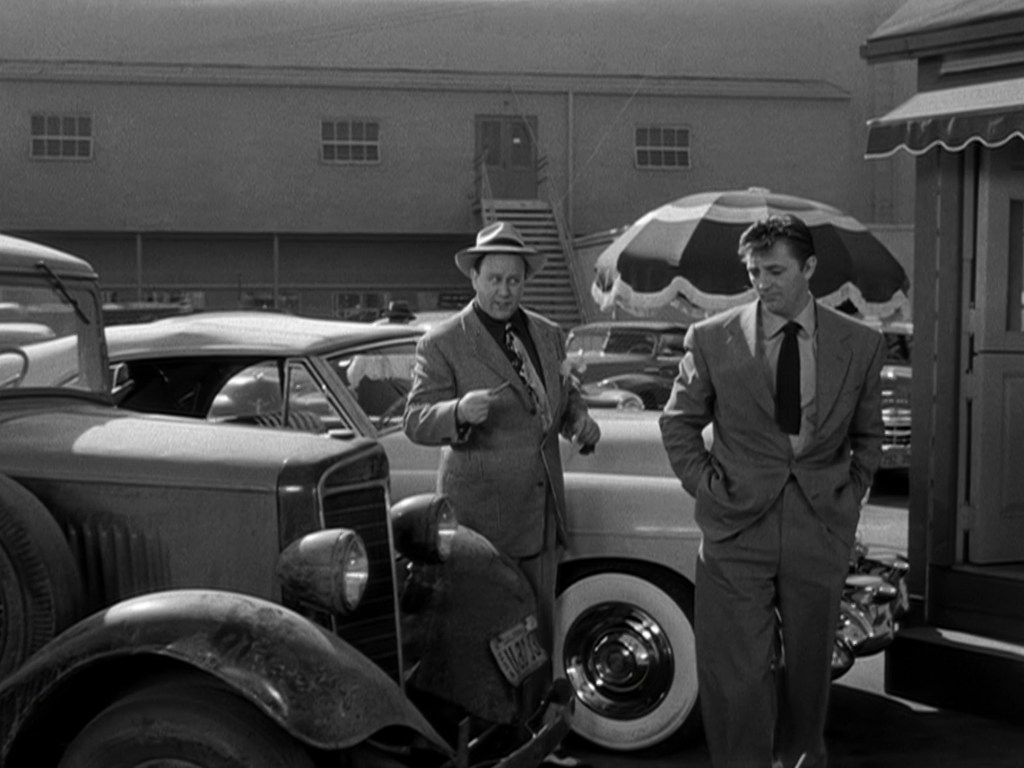 Two men in 1940s attire stand next to classic cars in a parking lot. One man gestures while speaking, and the other listens, both engaged in conversation.