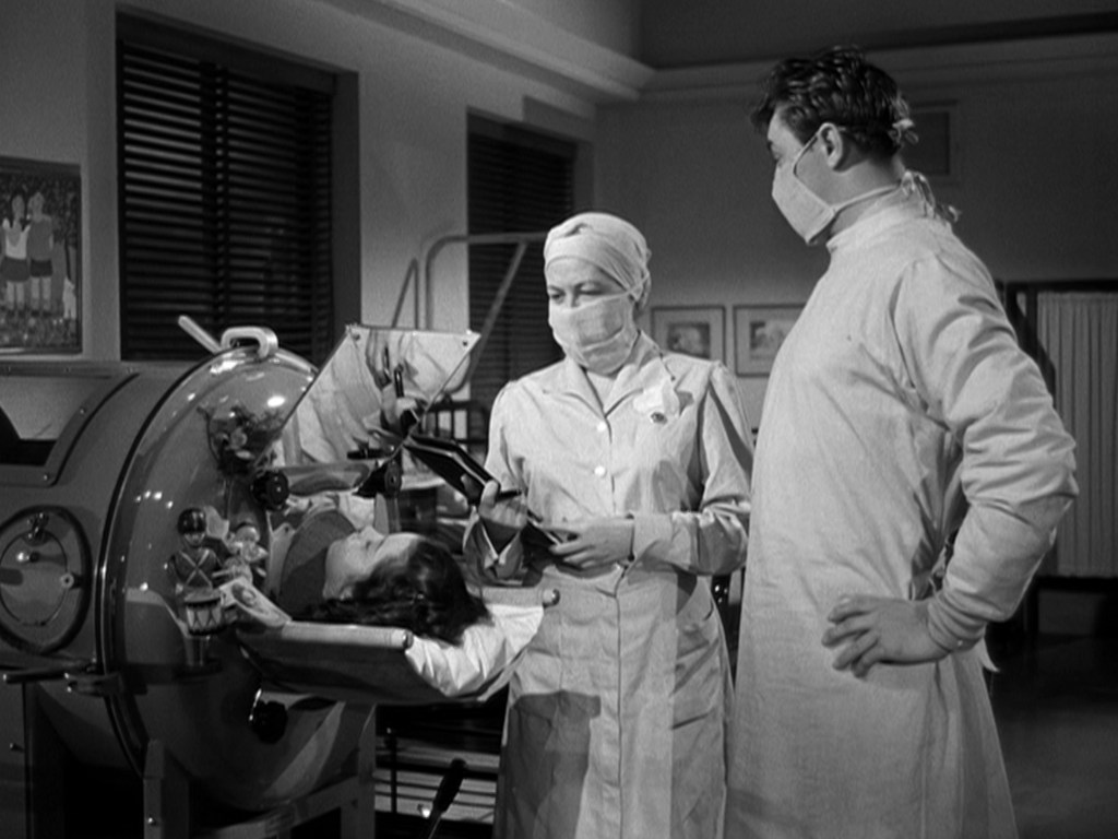 A nurse and a doctor wearing masks stand beside a patient in a medical device, likely a respirator, during a procedure in a hospital setting.