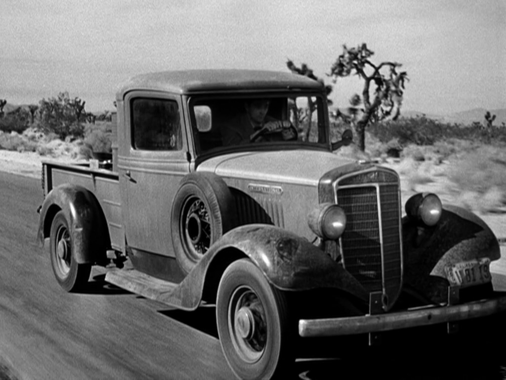 A vintage pickup truck driving on a barren road with desert scenery in the background.