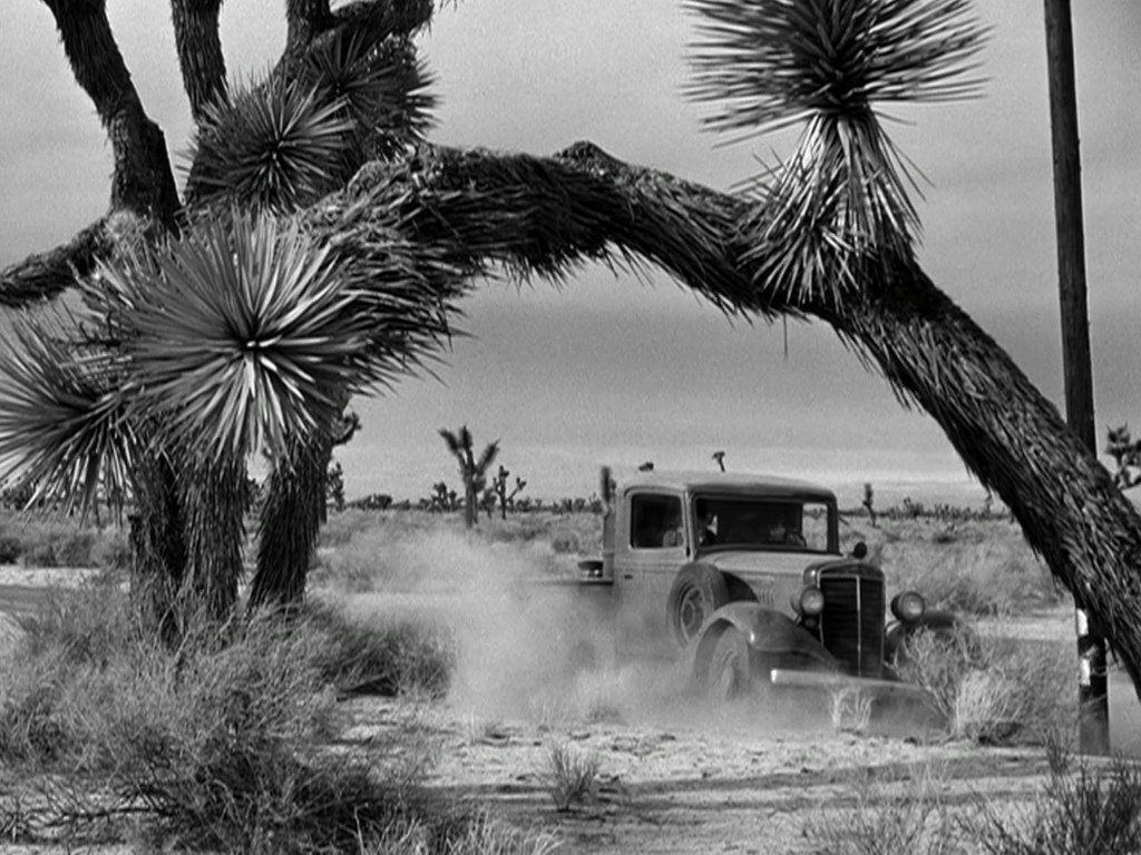 A vintage truck driving through a dusty desert landscape, framed by spiky desert plants under a cloudy sky.