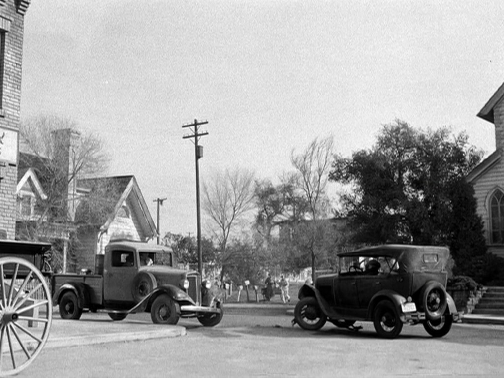 A vintage black and white scene featuring two classic cars, one a pickup truck and the other a convertible, near a street corner with old buildings and trees in the background.