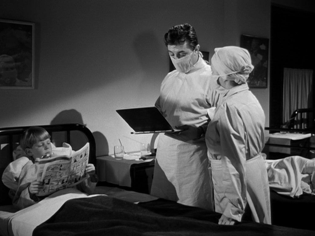 A young boy reading a comic book in a hospital bed while two medical staff, a man and a woman, discuss information from a laptop, all are wearing masks.