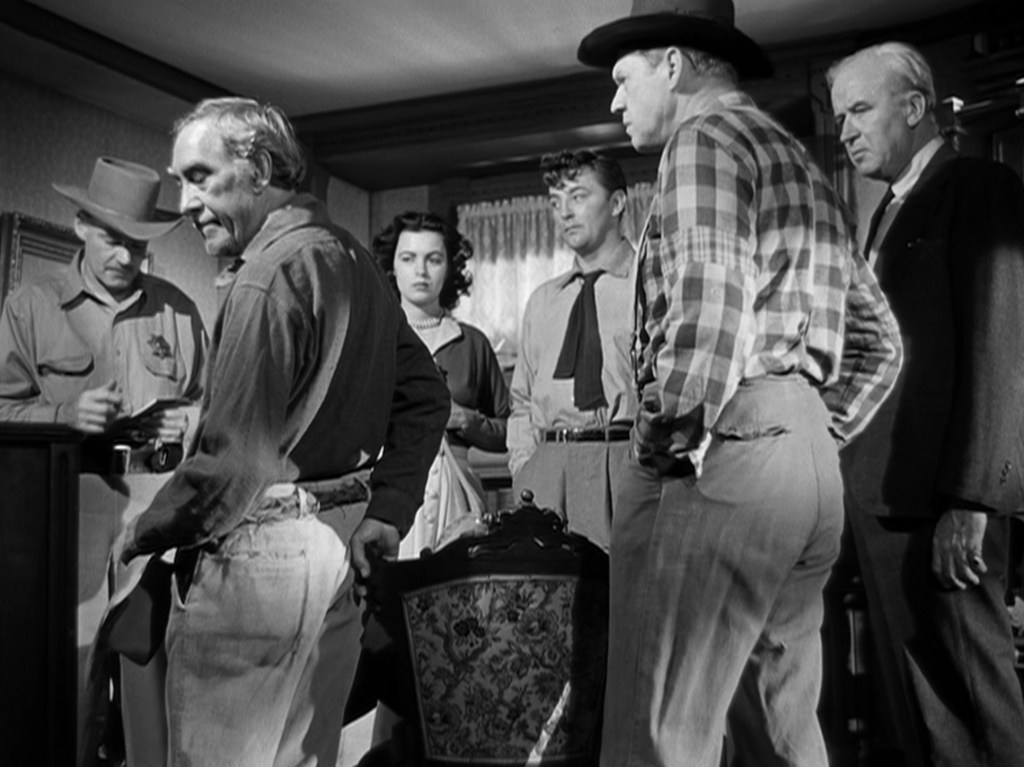 A group of six people in a dimly lit room, four men and two women, engaged in a serious discussion. One man is holding a clipboard while another appears to be speaking to the group. The setting includes a decorative chair in the foreground.