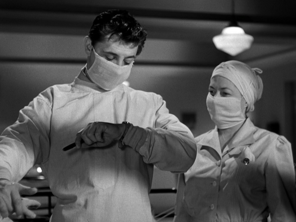 A male surgeon in a surgical mask checks his watch, while a female nurse in a headscarf observes him, both in a hospital setting.