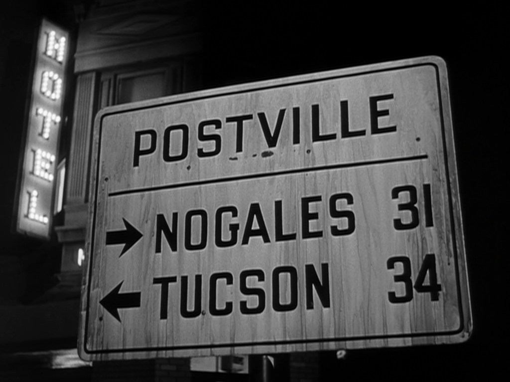 A weathered road sign indicating directions to Postville, Nogales (31 miles) and Tucson (34 miles), with a neon-lit motel sign in the background.