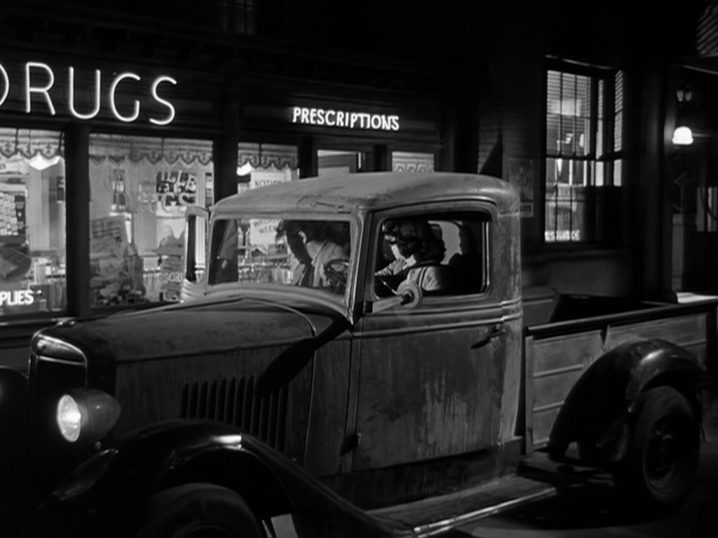 A vintage pickup truck parked in front of a dimly lit pharmacy with a neon sign reading 'DRUGS' and a window displaying various products.