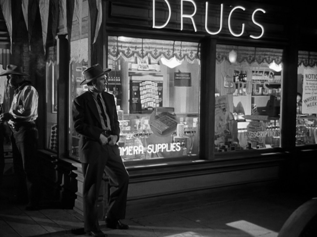 Black and white image of a man in a suit and hat leaning against a storefront labeled 'DRUGS', with various items displayed in the window.