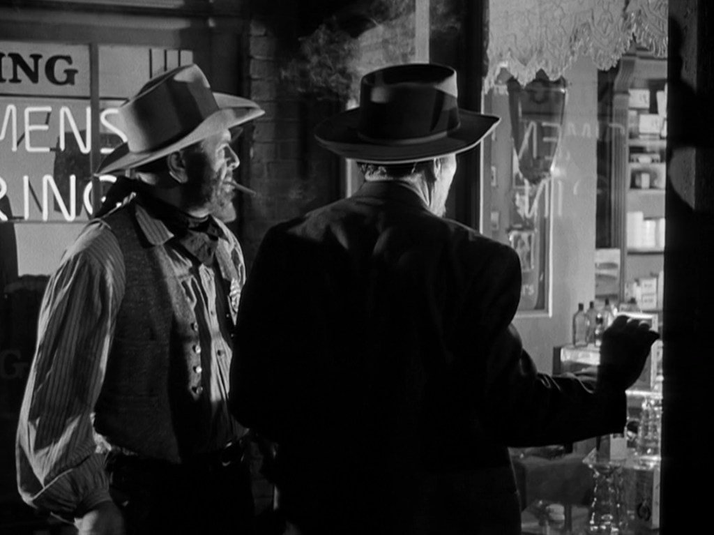 Two men in western attire, one with a mustache and a cigar, stand in front of a shop window that reads 'Men's Wearing'. The scene is in black and white, creating a vintage atmosphere.