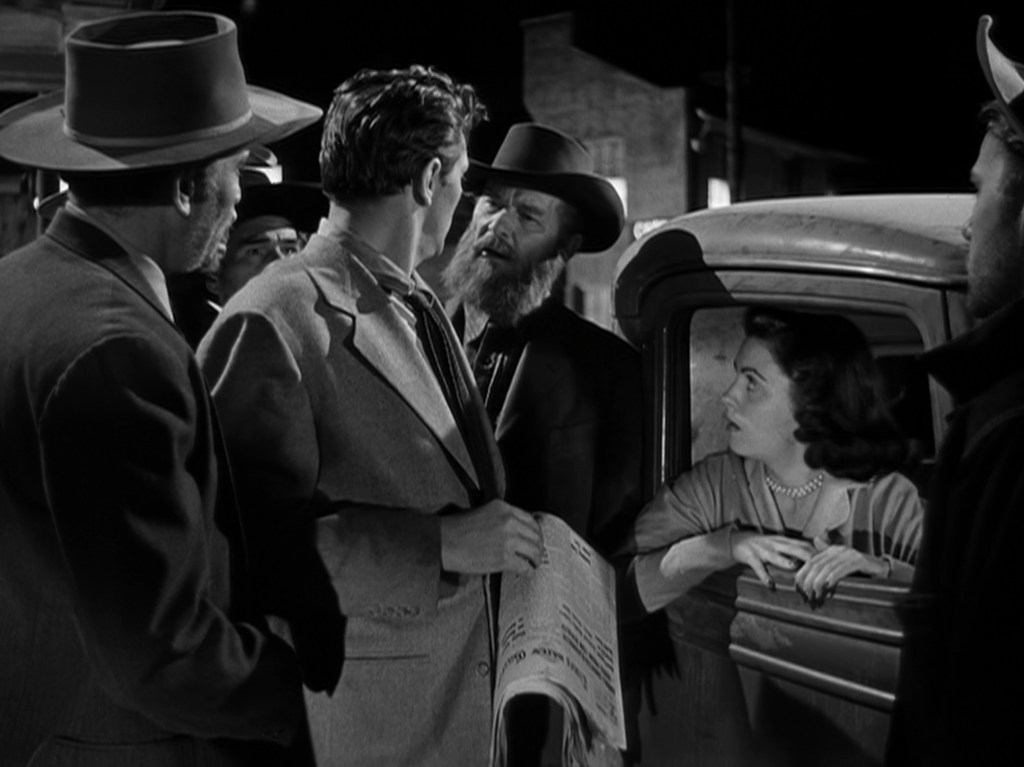 A tense scene featuring several men in hats confronting a man in a light-colored suit, while a woman in a car looks on with concern. They are surrounded by buildings and a night sky.