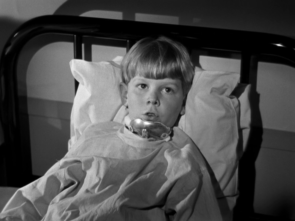 A young boy lying in a hospital bed, looking directly at the camera with a serious expression. He is wearing a hospital gown and there is a medical device attached to his mouth.