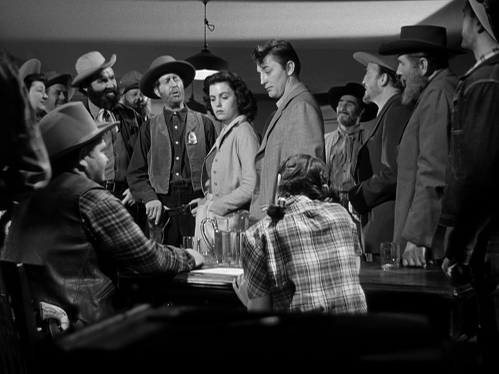 A tense scene in a western saloon with a group of cowboys and townspeople surrounding a man and a woman, engaging in a serious discussion.