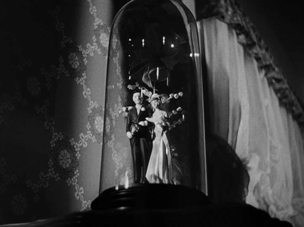 A close-up of a decorative glass dome enclosing a miniature bride and groom figurine, set against a patterned wallpaper backdrop.