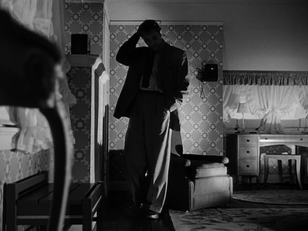 A man in a suit stands in a dimly lit room, looking pensively with one hand on his head, surrounded by vintage furniture and patterned wallpaper.