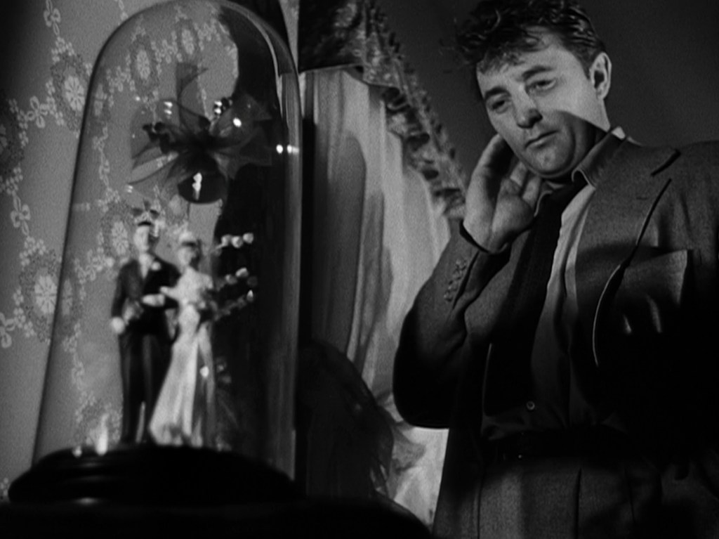 A man in a suit gazes thoughtfully at a decorative glass dome containing miniature wedding figurines, set against a backdrop of patterned wallpaper.