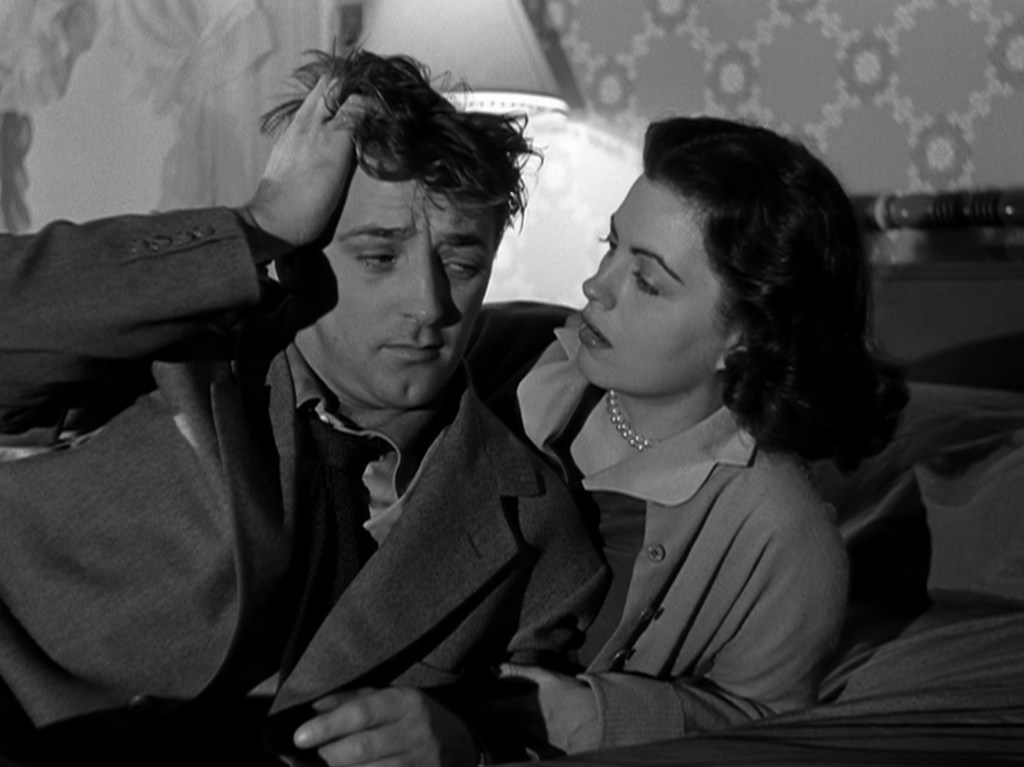 A man with tousled hair looks distressed while sitting on a bed, holding his head, as a woman leans in closely, showing concern.