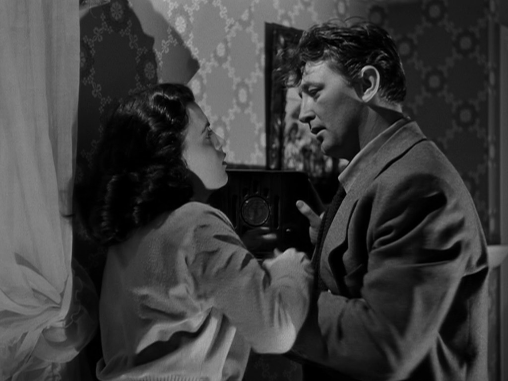 A black and white scene depicting a man and woman in close proximity, engaged in an intense conversation. The man is wearing a suit, while the woman is in a light sweater. They are positioned near a vintage radio against a patterned wall.