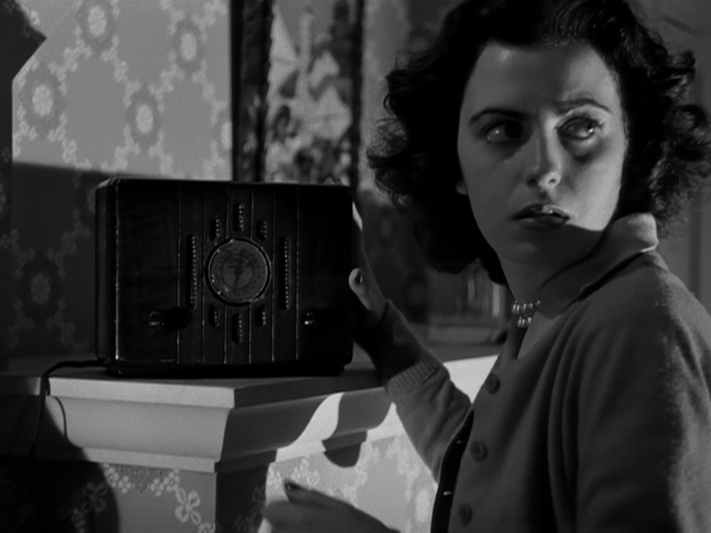 A woman with short dark hair looks anxiously over her shoulder while standing next to an old-fashioned radio in a dimly lit room.