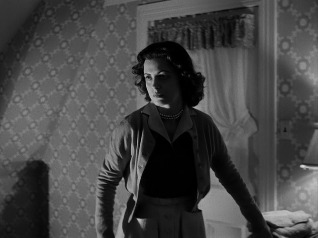 A woman with dark, curly hair stands in a dimly lit room, wearing a cardigan and a pearl necklace, looking thoughtfully towards the right. The room features patterned wallpaper and a window with curtains.