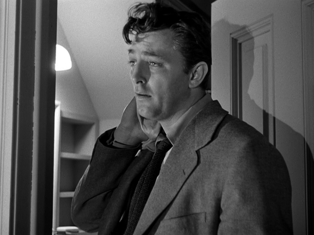 A black-and-white image of a man in a suit with tousled hair, looking thoughtful or troubled, standing in a doorway.