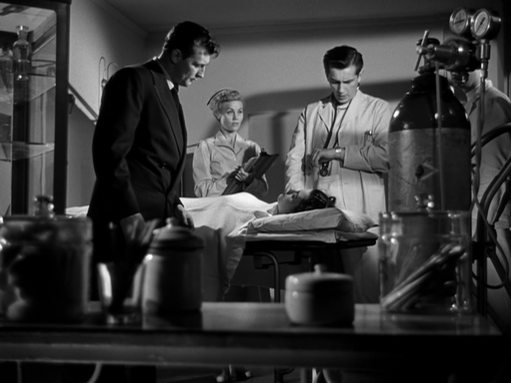 A black and white scene in a hospital setting, featuring three characters: two men in suits and lab coats observing a patient on a table, while a woman takes notes.