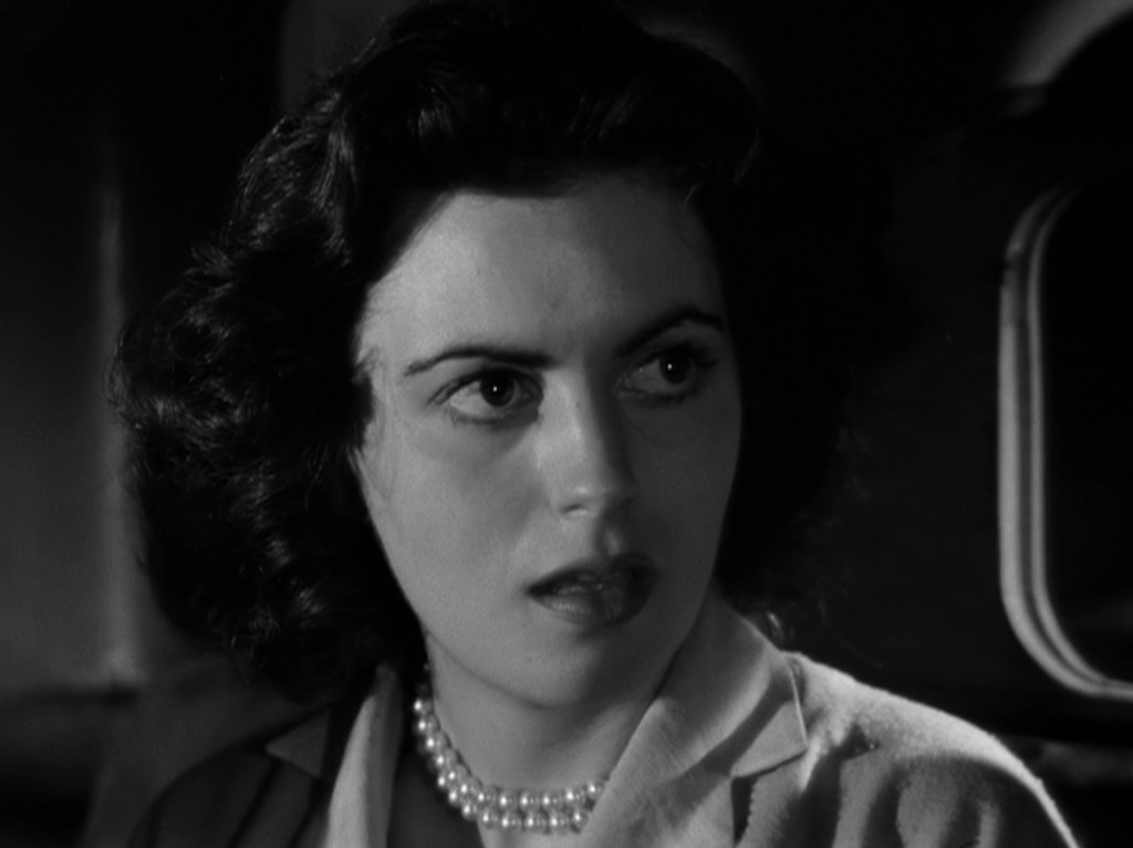 Close-up of a woman with dark, wavy hair and a pearl necklace, looking concerned or surprised in a black-and-white scene.